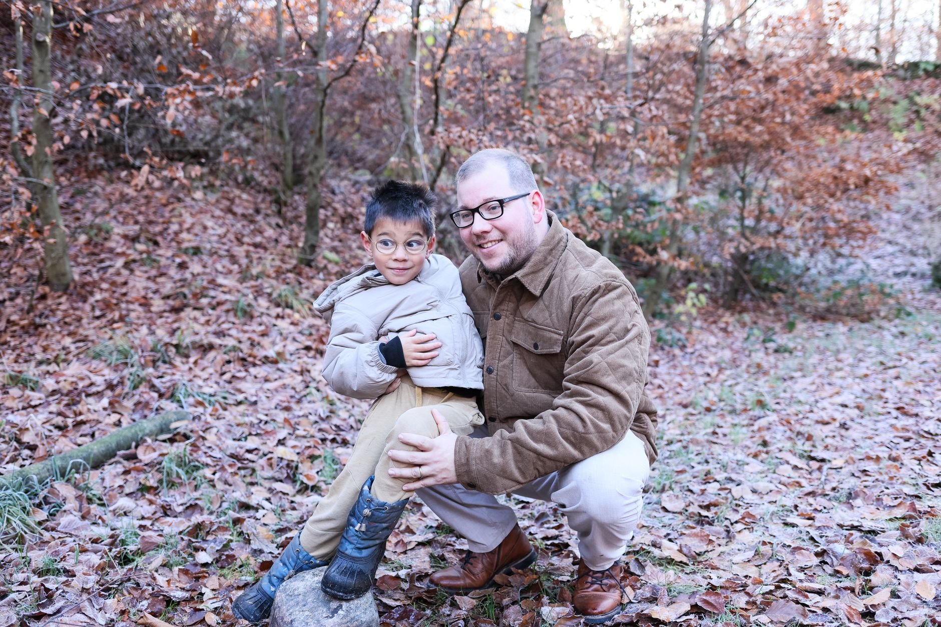 A father and son share a joyful moment in a fall forest setting, surrounded by autumn leaves.