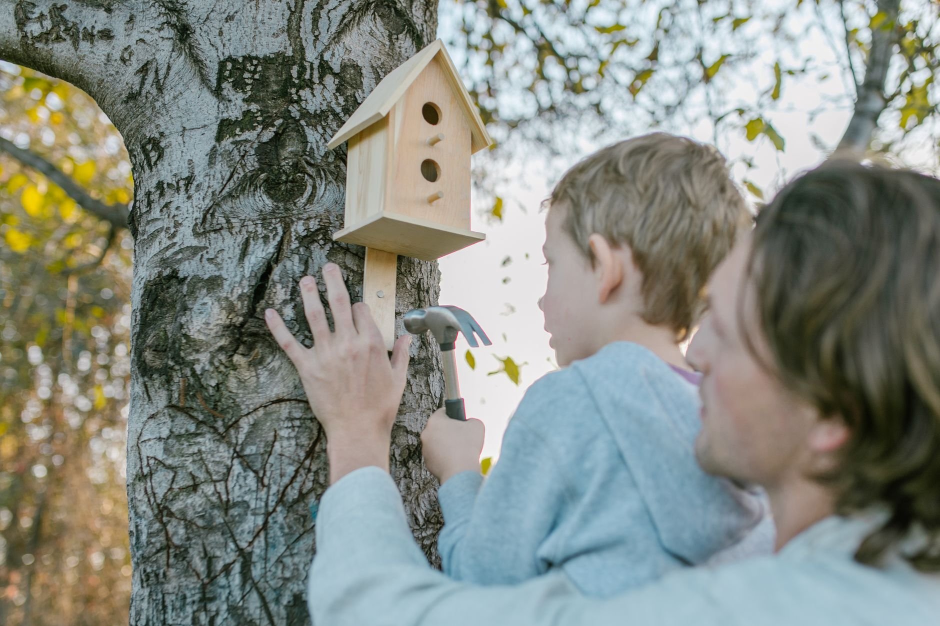 Father helps son mount a wooden birdhouse on a tree using a hammer.